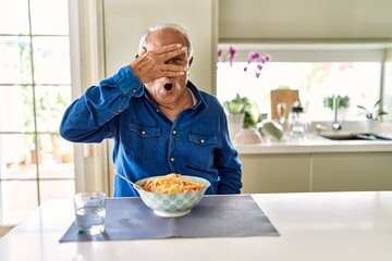 Senior man with grey hair eating pasta spaghetti at home peeking in shock covering face and eyes with hand, looking through fingers with embarrassed expression.