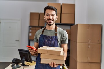 Wall Mural - Young hispanic man business worker using smartphone holding packages at storehouse