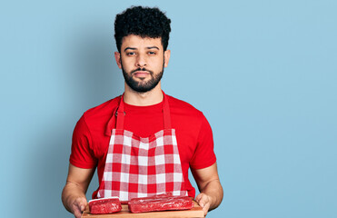 Poster - Young arab man with beard holding board with raw meat relaxed with serious expression on face. simple and natural looking at the camera.