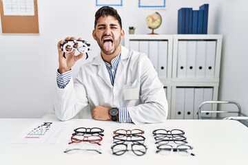 Poster - Young optician man holding optometry glasses sticking tongue out happy with funny expression. emotion concept.