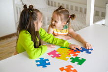 Funny Girls Collect Puzzles Together At The Table In The Children's Room. Large Bright Colorful Puzzle Pieces Are A Symbol Of Autism. Communication And Relationship Between Kids