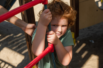 Wall Mural - curly boy near rope ladder and mothers hand in amusement park.