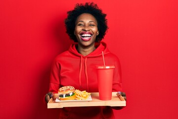 Wall Mural - Young african american woman eating a tasty classic burger with fries and soda smiling and laughing hard out loud because funny crazy joke.