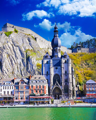 Dinant, Belgium - March 9. 2022: View over river meuse on picturesque series of old colorful houses, rock wall with citadel, gothic church against clear blue winter sky, fluffy clouds