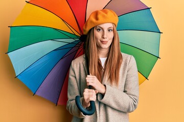 Young irish woman wearing french style holding colorful umbrella thinking attitude and sober expression looking self confident