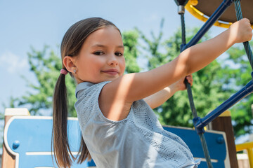 Wall Mural - girl smiling at camera while climbing on rope ladder on playground.