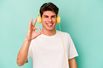 Young caucasian man listening to music isolated on blue background cheerful and confident showing ok gesture.