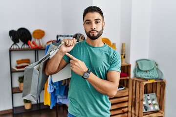 Sticker - Young handsome man with beard holding shopping bags at retail shop pointing with hand finger to the side showing advertisement, serious and calm face