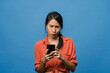 © tirachard - Thinking dreaming young Asia lady using phone with positive expression, dressed in casual clothing feeling happiness and stand isolated on blue background. Happy adorable glad woman rejoices success.