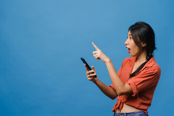 Wall Mural - Portrait of young Asian lady using mobile phone with cheerful expression, show something amazing at blank space in casual clothing and stand isolated over blue background. Facial expression concept.