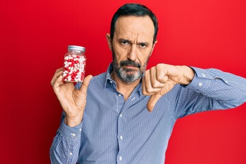 Poster - Middle age hispanic man holding jar of pills with angry face, negative sign showing dislike with thumbs down, rejection concept