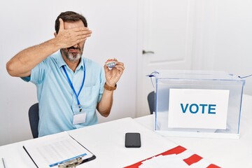 Canvas Print - Middle age man with beard sitting by ballot holding i vote badge covering eyes with hand, looking serious and sad. sightless, hiding and rejection concept