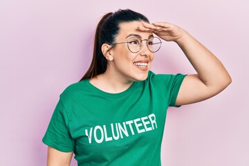 Sticker - Young hispanic woman wearing volunteer t shirt very happy and smiling looking far away with hand over head. searching concept.