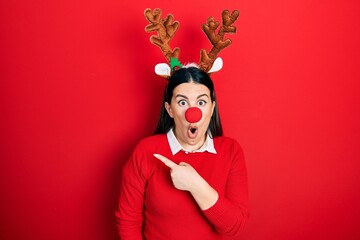 Poster - Young hispanic woman wearing deer christmas hat and red nose surprised pointing with finger to the side, open mouth amazed expression.