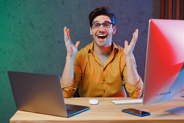 Young shocked amazed excited surprised employee programmer IT specialist business man in casual shirt sit work at office desk with pc laptop desktop computer spread hands Achievement career concept.