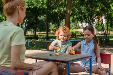 Wall Mural - woman sitting near children playing with toys and sand in park.