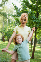 Wall Mural - excited woman holding hands of son while having fun in summer park.