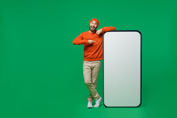 Wall Mural - Full body young smiling fun man 20s wear orange sweatshirt hat point index finger on big blank screen mobile cell phone with workspace copy space mockup area isolated on plain green background studio.