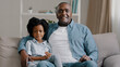 © Yuliia - African american family sitting on sofa kid girl waving hello greeting adult father hugging daughter talking at camera smiling showing healthy white teeth after dental procedure video chat webcam view