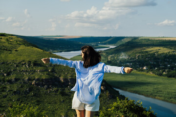 Wall Mural - Freedom stylish girl with hands up standing on the hill with the river and hills on the background. Summer travel.