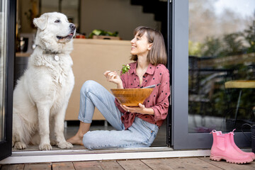 Happy woman eats salad while sitting with her adorable huge white dog on terrace near the window. Concept of friendship with pets and healthy eating