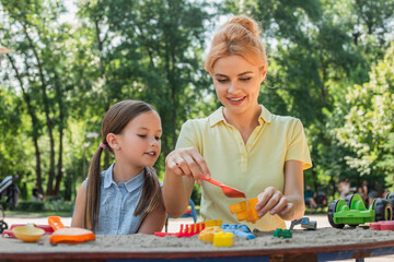 Wall Mural - smiling woman playing with toy shovel and sand near daughter.