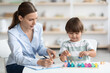 © Prostock-studio - Cute little boy playing colorful wooden game at specialist office, professional woman therapist taking notes, free space