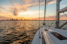 Yacht Sailing Towards Cityscape On Horizon At Sunset
