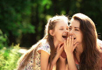 Wall Mural - Happy enjoying mother speaking and laughing with her relaxing smiling kid girl on bright summer green trees background. Closeup