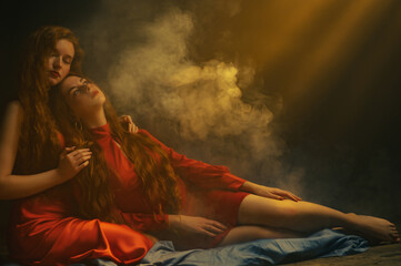 Studio art portrait of two beautiful redhead freckled women wearing red dresses posing in smoke, darkness and warm light. Copy, empty space for text