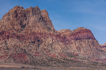 Wall Mural - Las Vegas, Nevada, USA - February 23, 2010: Red Rock Canyon Conservation Area. Tall rocky mountain has layer of red rock cutting through it halfway its height under blue sky. Desert floor up front.