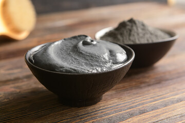 Bowls of activated carbon facial mask and powder on wooden table, closeup