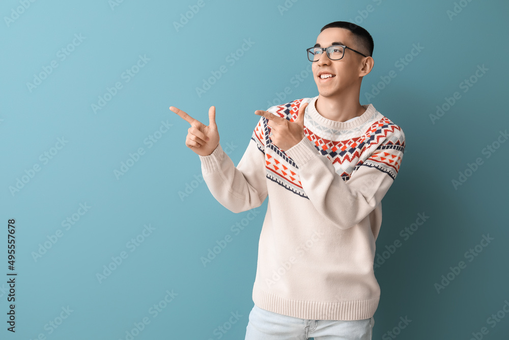 Handsome young Asian man in knitted sweater pointing at something on blue background
