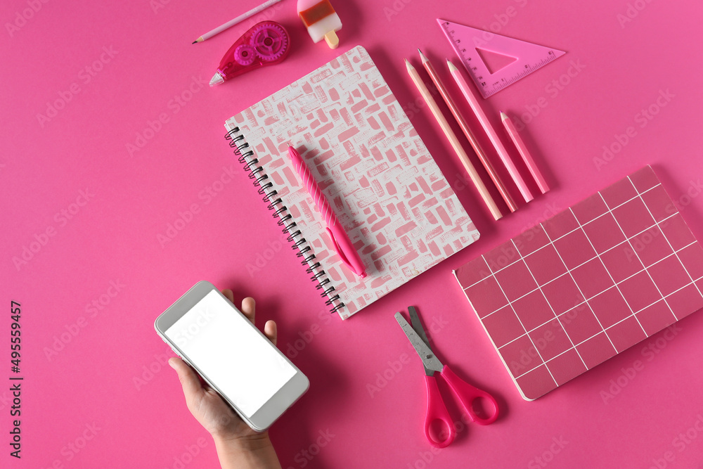 Child's hand with mobile phone and different stationery on pink background