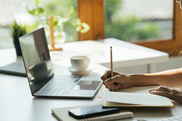 Woman's hands on laptop keyboard. She is also taking notes. coffee cup and white binders are standing on the desk.