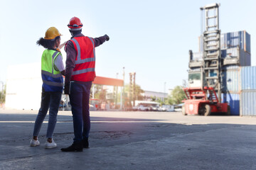 Behind two industrial African American engineer man and woman wearing safety vest and helmet, pointing and looking at container lifting truck, workers working at logistic shipping cargo yard workplace