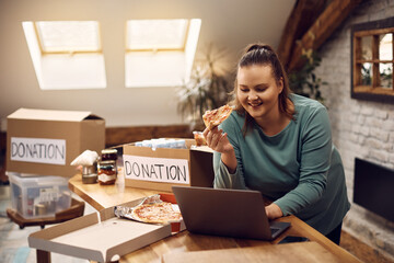 Charity community worker works on laptop and eats pizza while organizing donation boxes.