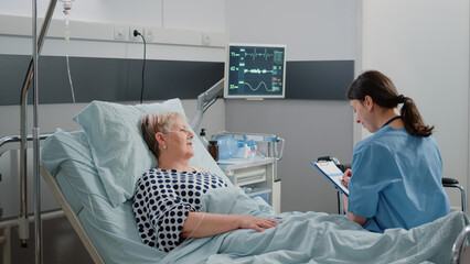 Wall Mural - Medical assistant doing consultation with retired patient in hospital ward bed. Nurse talking to ill woman for healthcare treatment and disease, giving assistance and support for recovery