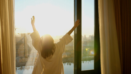 Young woman in white bathrobe open curtains in bedroom after waking up. Lady stretching with rising hands, looking out panoramic window enjoying sunrise in early morning