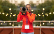 © Syda Productions - people, profession and photography concept - happy young woman or press photographer with digital camera and conference id card photographing over garland lights on roof top party background