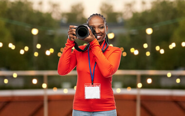 Sticker - people, profession and photography concept - happy young woman or press photographer with digital camera and conference id card photographing over garland lights on roof top party background