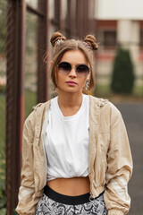 Wall Mural - Stylish beauty girl with hairstyle, pink lips and round vintage sunglasses in sporty clothes with windbreaker, t-shirt, and leggings walks on the street .