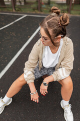 Wall Mural - Beautiful street woman with hairstyle and sunglasses in fashion sports clothes with jacket, t-shirt, shorts and sneakers sits and poses outdoors