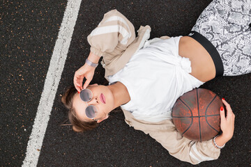 Wall Mural - Fashion young beautiful woman with vintage sunglasses in stylish clothes with ball lies and rest on basketball court, top view. Trendy cool hipster girl on asphalt