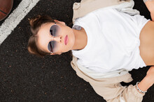 Beautiful Stylish Woman Model With Vintage Sunglasses And Hairstyle In A Fashionable Windbreaker And White T-shirt Lies And Rest On The Pavement, Top View. Trendy Female Portrait. Fashion And Beauty
