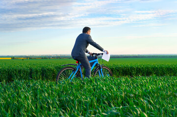 businessman dressed in a business suit, rides a bicycle through a green grass field, he has a briefcase and documents, beautiful nature in spring, business concept