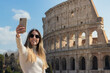 Happy young woman taking a selfie in front of the colosseum while in Rome, Italy