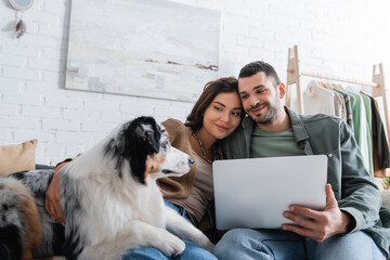 Wall Mural - pleased young couple looking at australian shepherd dog near laptop.