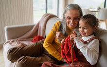 Little Girl Sitting On Sofa With Her Grandmother And Learning To Knit Indoors At Home.