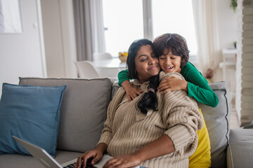 indian mother using laptop and her little son hugging herg on sofa at home.